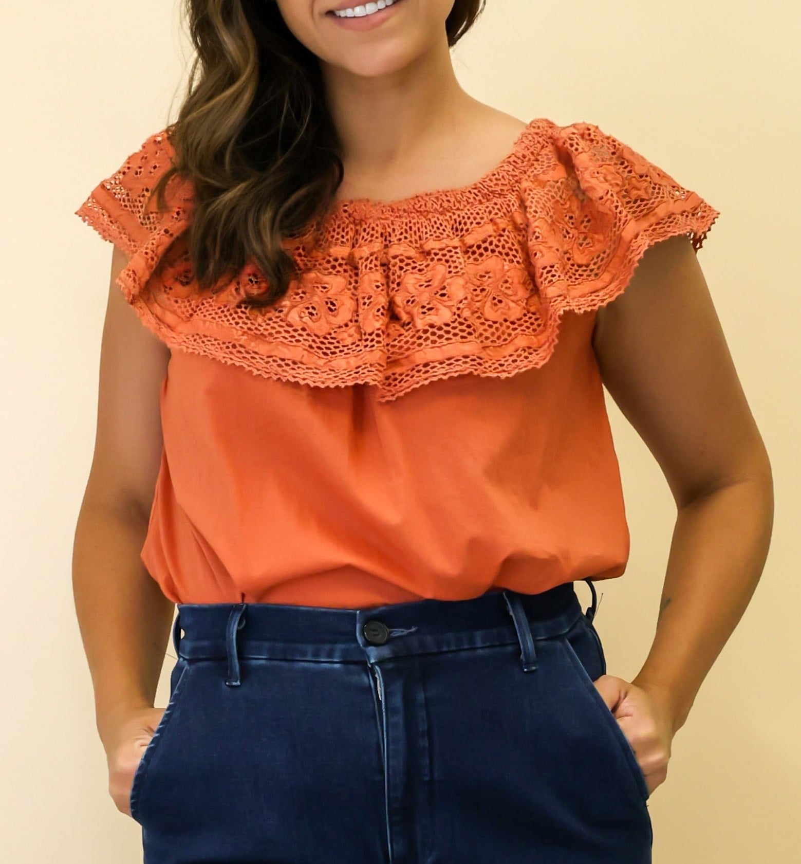 Woman wearing an orange top with ruffled sleeves and blue jeans on a beige background