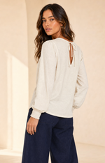 A woman wearing a cream-colored long-sleeve knit top with a tie-back detail, paired with dark navy wide-leg pants, shown from a three-quarters back view in a warm, minimalist studio setting.
