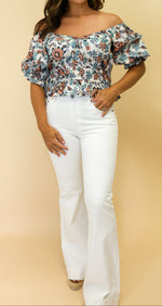 Woman wearing a floral off-shoulder top and white pants against a beige background