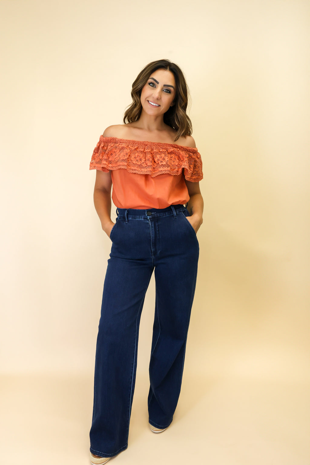 Woman wearing an orange off-shoulder top and blue jeans on a beige background
