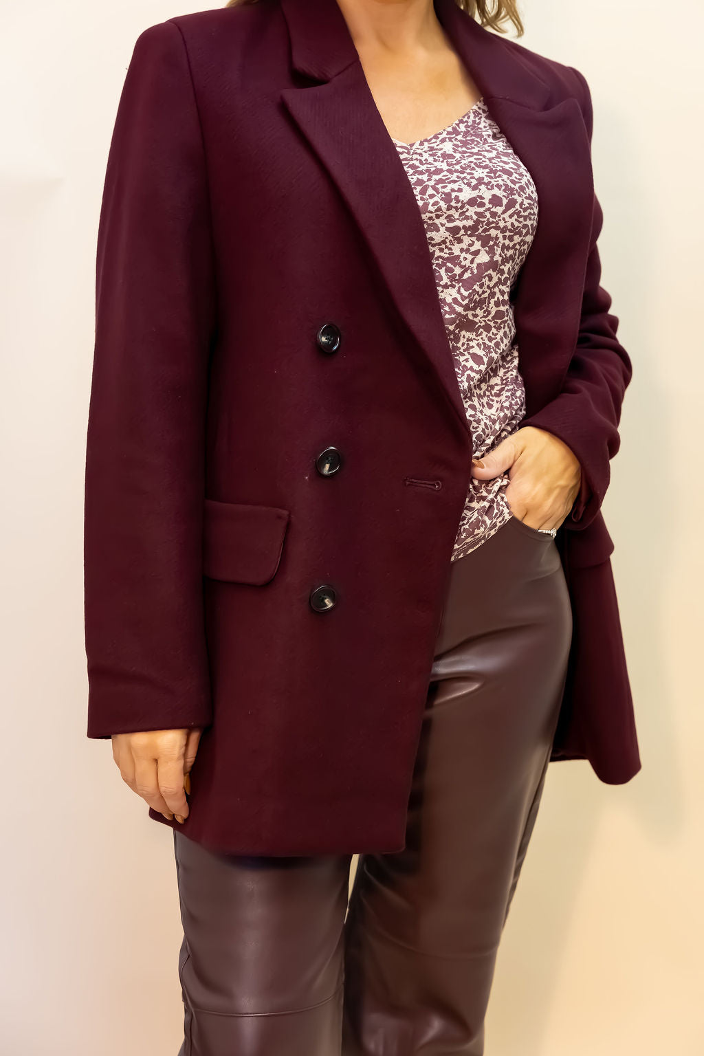 Person wearing a burgundy blazer over a patterned top and brown pants on a plain background