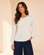 A woman modeling a cream-colored long-sleeve knit top with gathered cuffs, paired with dark navy wide-leg pants, set against a warm beige architectural background.