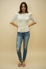 Person wearing a white ruched top and blue jeans against a beige background