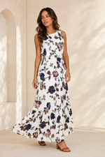 A woman modeling a sleeveless white maxi dress with a dark floral print, standing in a warm, sunlit architectural setting with cream-colored walls and arches.