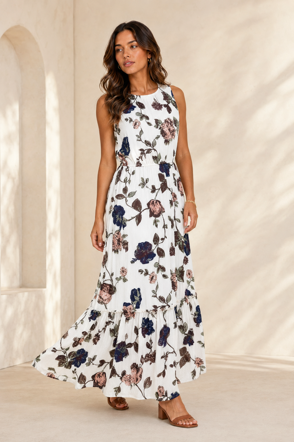 A woman modeling a sleeveless white maxi dress with a dark floral print, standing in a warm, sunlit architectural setting with cream-colored walls and arches.